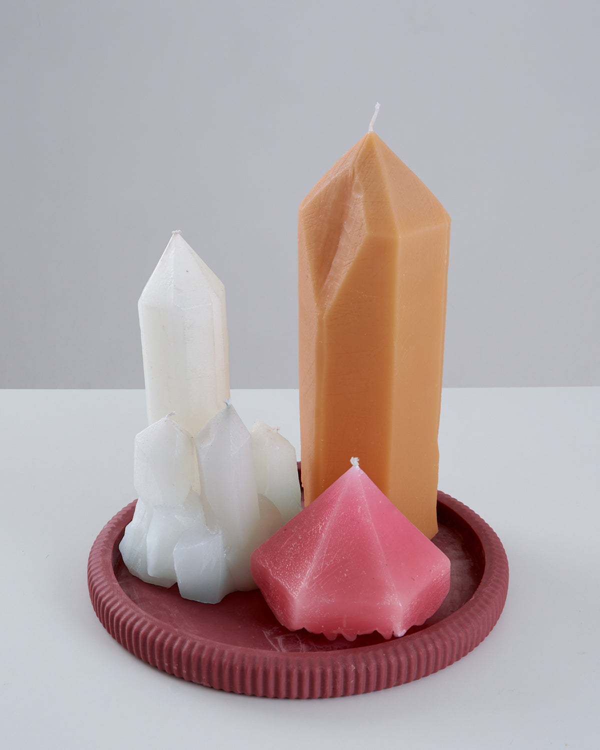 Três velas artesanais em formato de cristal reunidas sobre um prato/suporte redondo estriado na cor vinho: a vela Citrino na cor caramelo em formato de ponta facetada alta, uma vela branca em formato de drusa de cristais agrupados e uma vela rosa em formato de diamante lapidado, sobre superfície branca com fundo claro