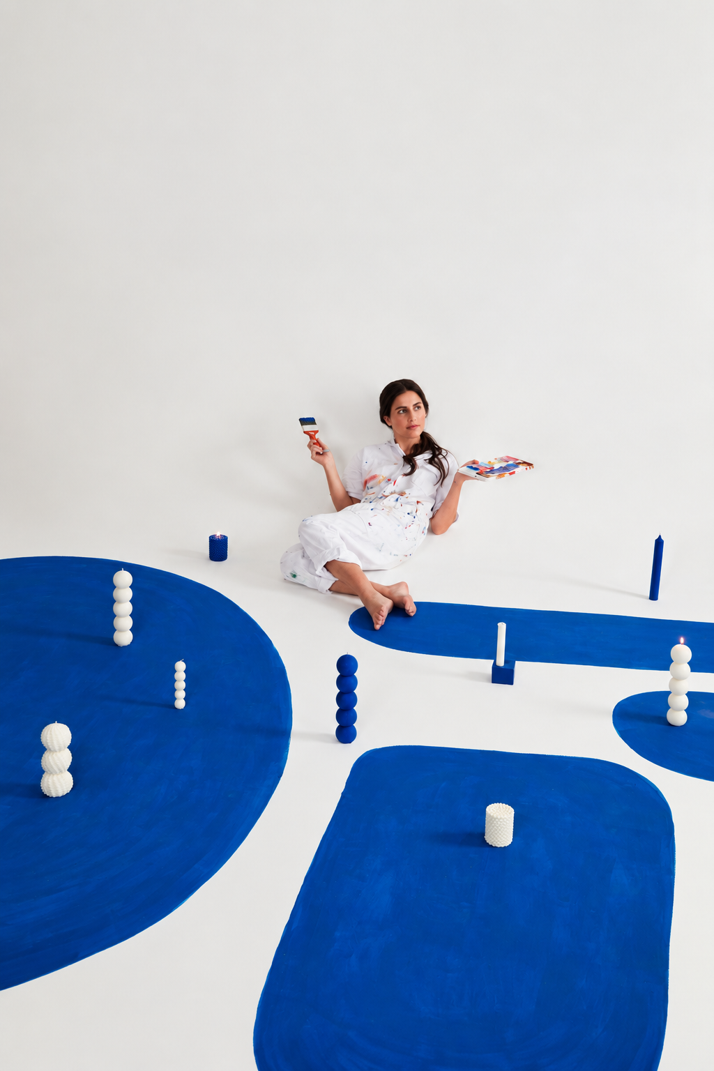 Diversas velas Bolas Grandes artesanais em formato de esferas empilhadas, nas cores branca e azul, distribuídas sobre formas orgânicas pintadas de azul no chão de um estúdio branco; ao centro, uma modelo em roupa branca com respingos de tinta, sentada segurando uma trincha e uma paleta de tintas, em cena editorial