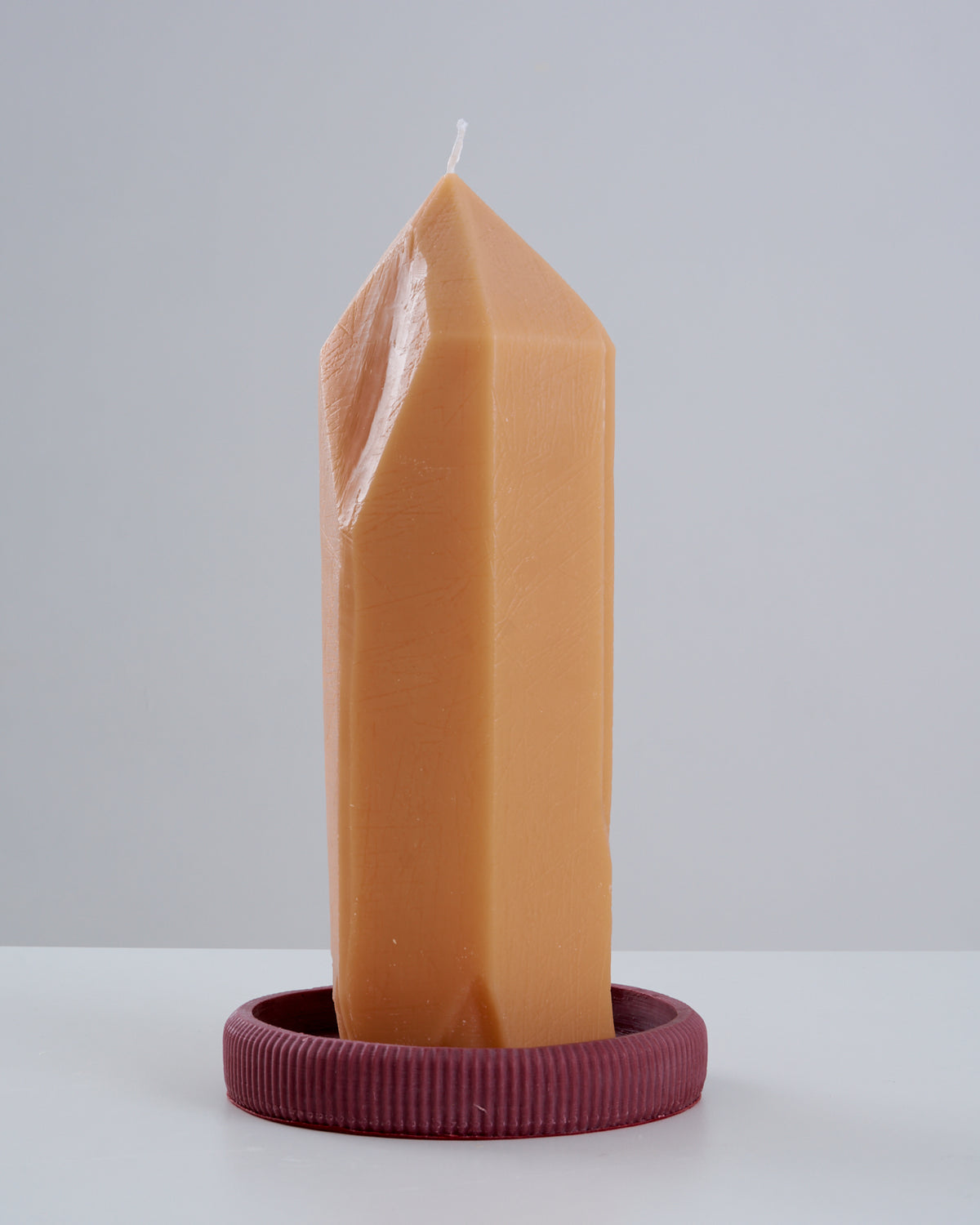 Vela Citrino artesanal na cor caramelo, em formato de ponta de cristal facetada com topo em bico, apoiada sobre um prato/suporte redondo estriado na cor vinho, isolada sobre superfície branca com fundo claro