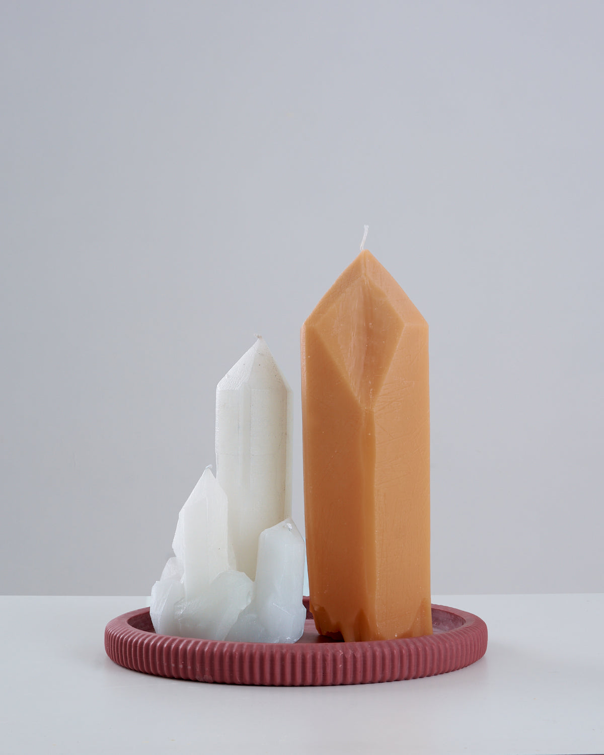 Vela Citrino artesanal na cor caramelo, em formato de ponta de cristal facetada, ao lado de uma vela branca em formato de drusa de cristais agrupados, ambas apoiadas sobre um prato/suporte redondo estriado na cor vinho, sobre superfície branca com fundo claro