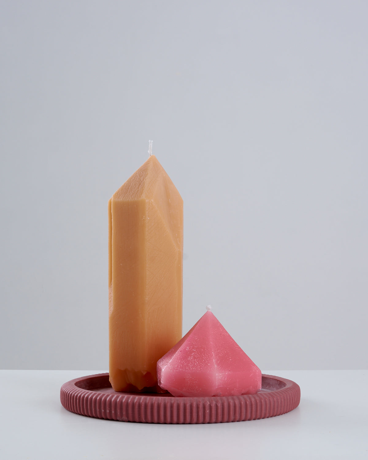 Vela Citrino artesanal na cor caramelo, em formato de ponta de cristal facetada, ao lado de uma vela rosa em formato de diamante lapidado de perfil baixo, ambas apoiadas sobre um prato/suporte redondo estriado na cor vinho, sobre superfície branca com fundo claro