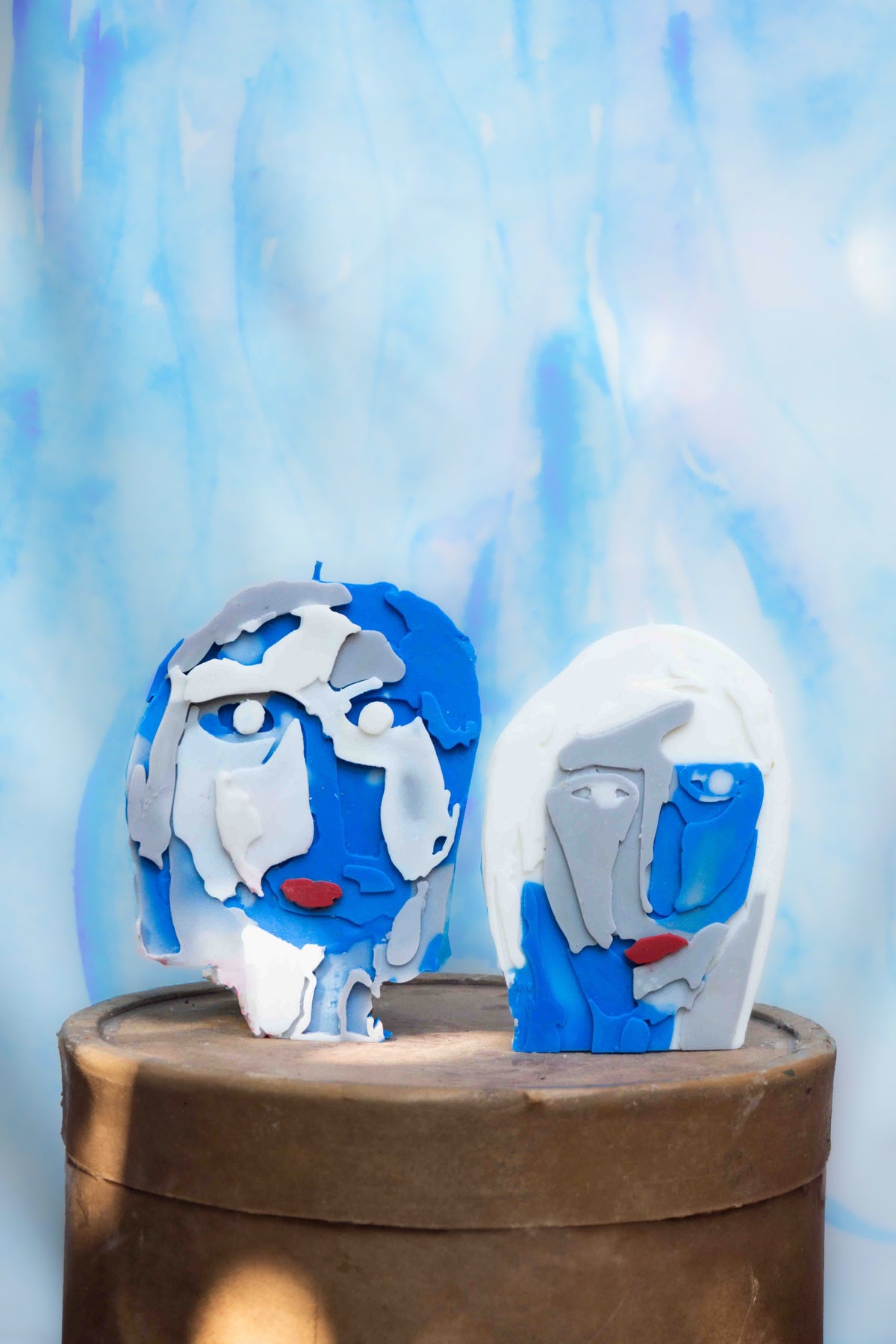 Duas velas La Foule Eleonore artesanais em formato de rosto humano estilizado, com relevo de feições em tons de azul, branco, cinza e boca vermelha, dispostas lado a lado sobre um pedestal cilíndrico de madeira, com fundo azul marmorizado